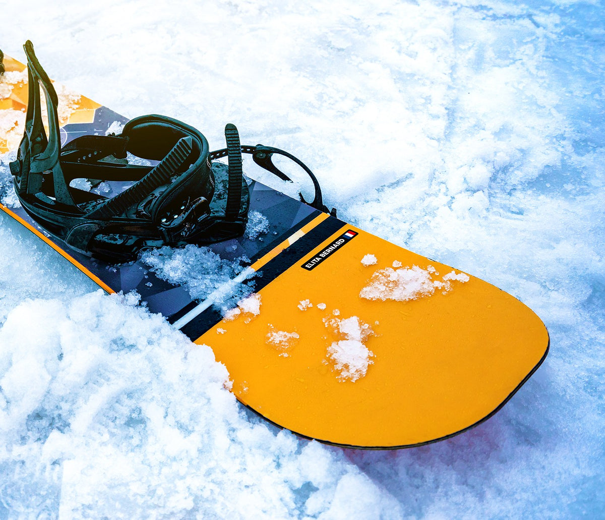 Geel snowboard met binding in de sneeuw, met een zwarte naamsticker met vlag op de plank